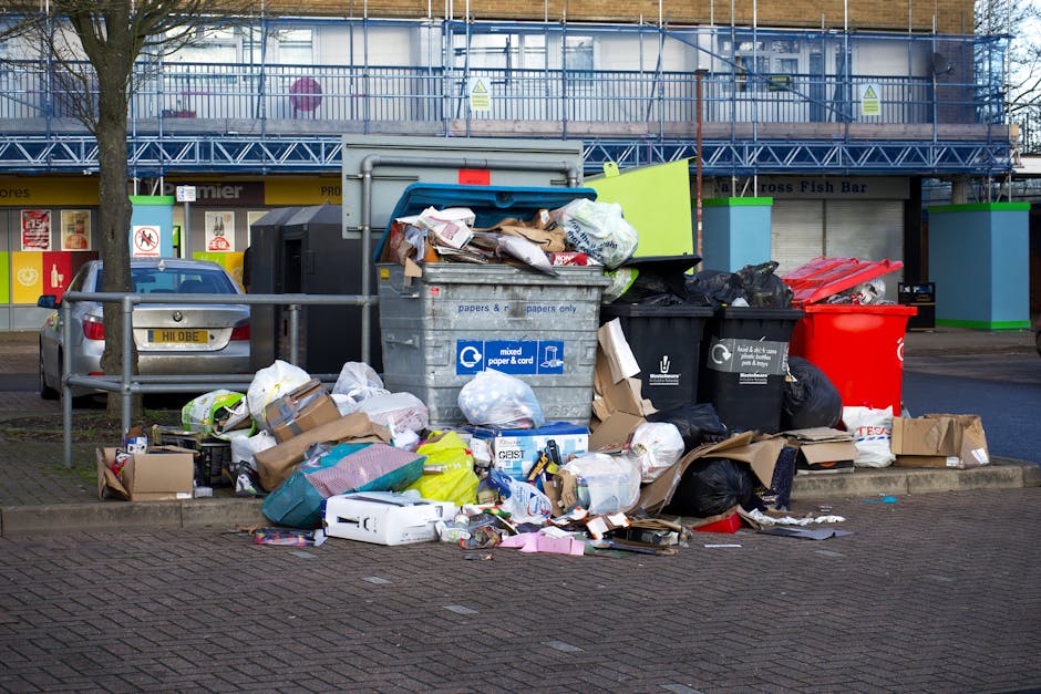 A collection of overflowing waste bins and discarded rubbish on a paved urban sidewalk, with various materials such as cardboard boxes, plastic bags, paper, and packaging boxes spilling onto the groun