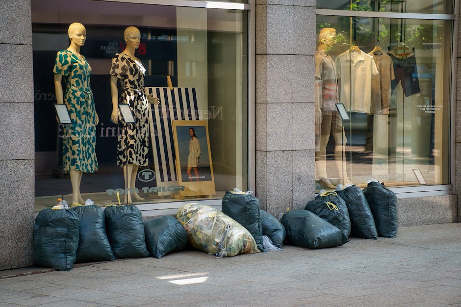 A collection of overflowing waste bins and discarded rubbish on a paved urban sidewalk, with various materials such as cardboard boxes, plastic bags, paper, and packaging boxes spilling onto the groun